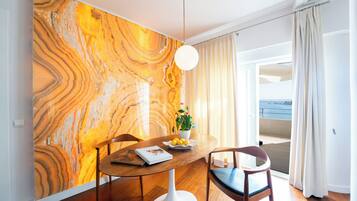 A dining area with a marble wall, a round table, two chairs, a potted plant, and a view of the sea through a balcony.