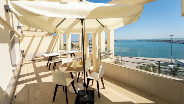 Un espace extérieur ensoleillé, aménagé avec une table et des chaises, un parasol blanc et une vue sur la mer.