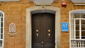 Une façade d’immeuble avec une porte à double battant de couleur sombre, une enseigne indiquant « Plaza Mina Suites » et une autre, bleue, indiquant « AT Ciudad ».