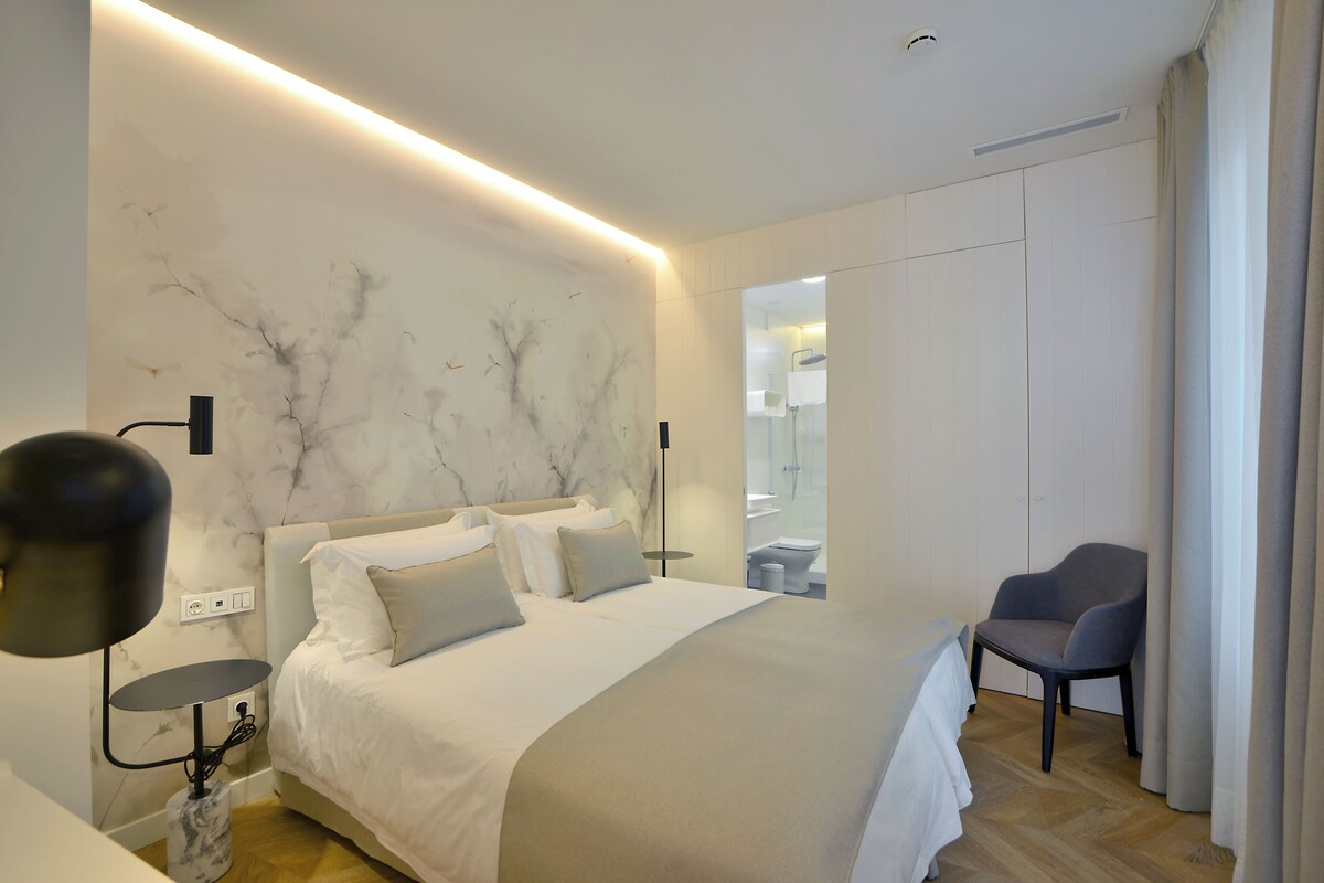 A modern hotel room with a large bed, a chair, a nightstand, and a marble wall.