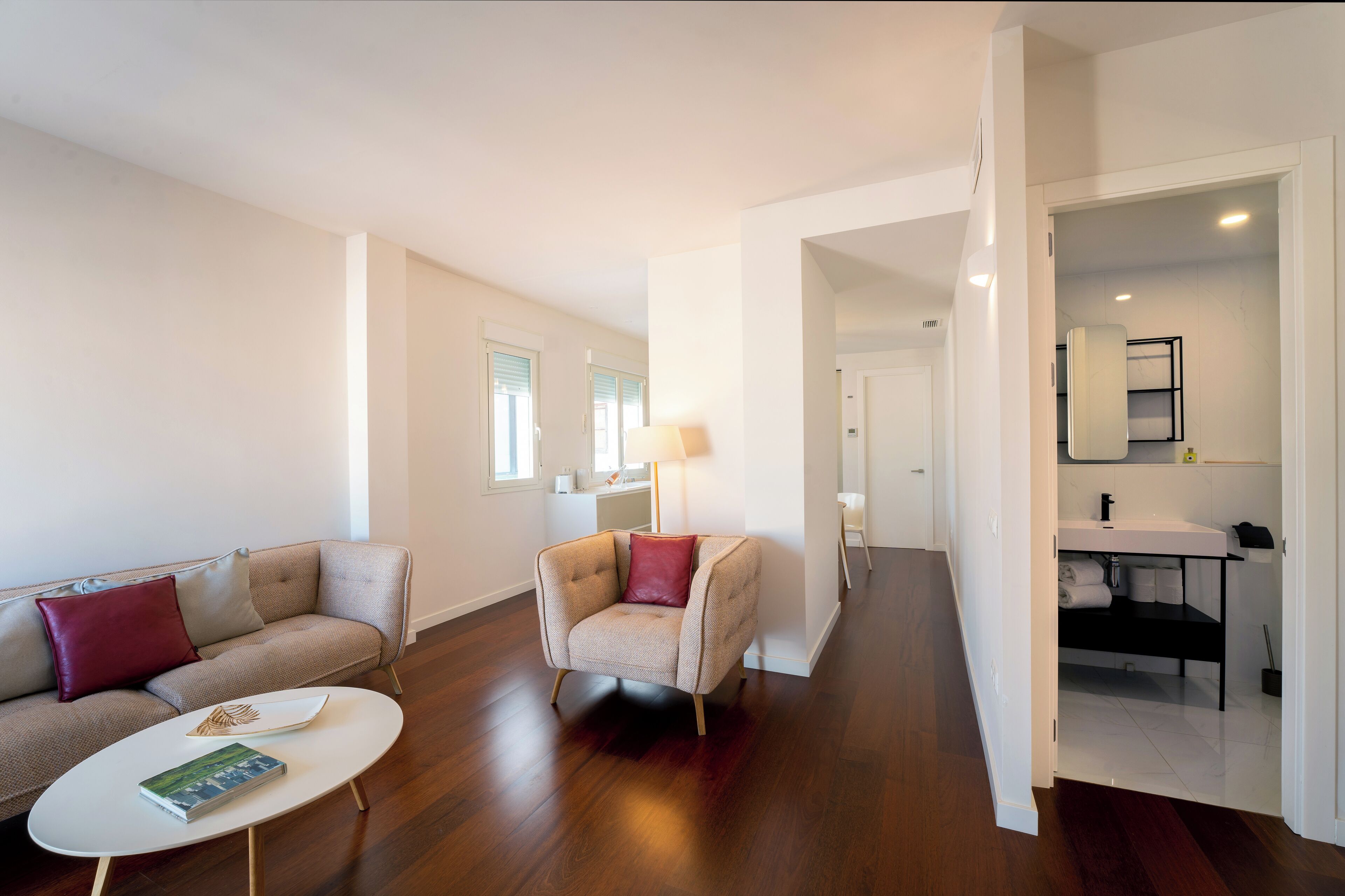 Una sala de estar moderna con un sofá, un sillón y una mesa de centro. Un pasillo conduce a un baño con un lavamanos y un espejo.