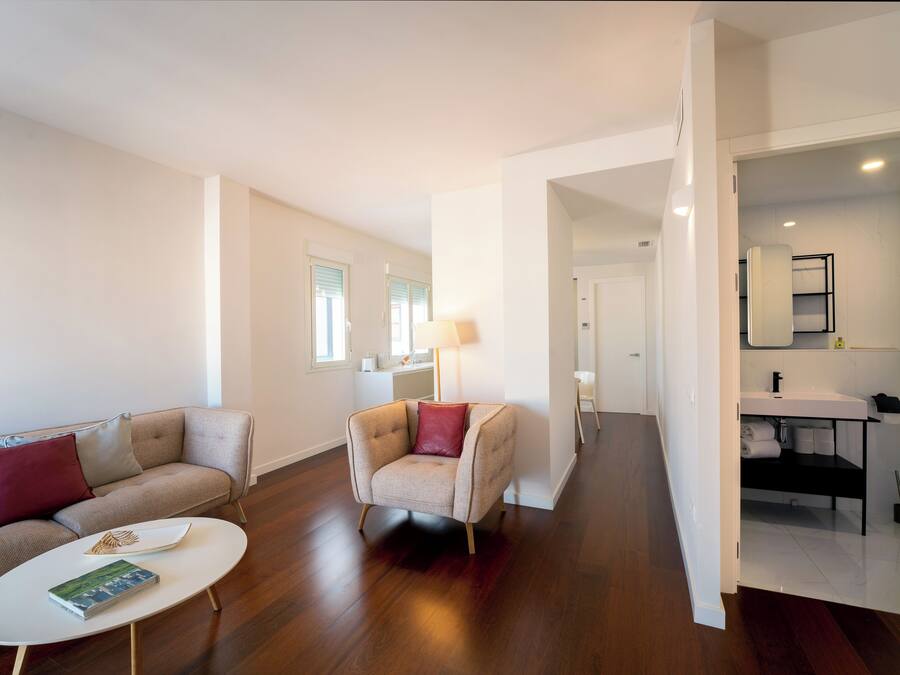 Una sala de estar moderna con un sofá, un sillón y una mesa de centro. Un pasillo conduce a un baño con un lavamanos y un espejo.