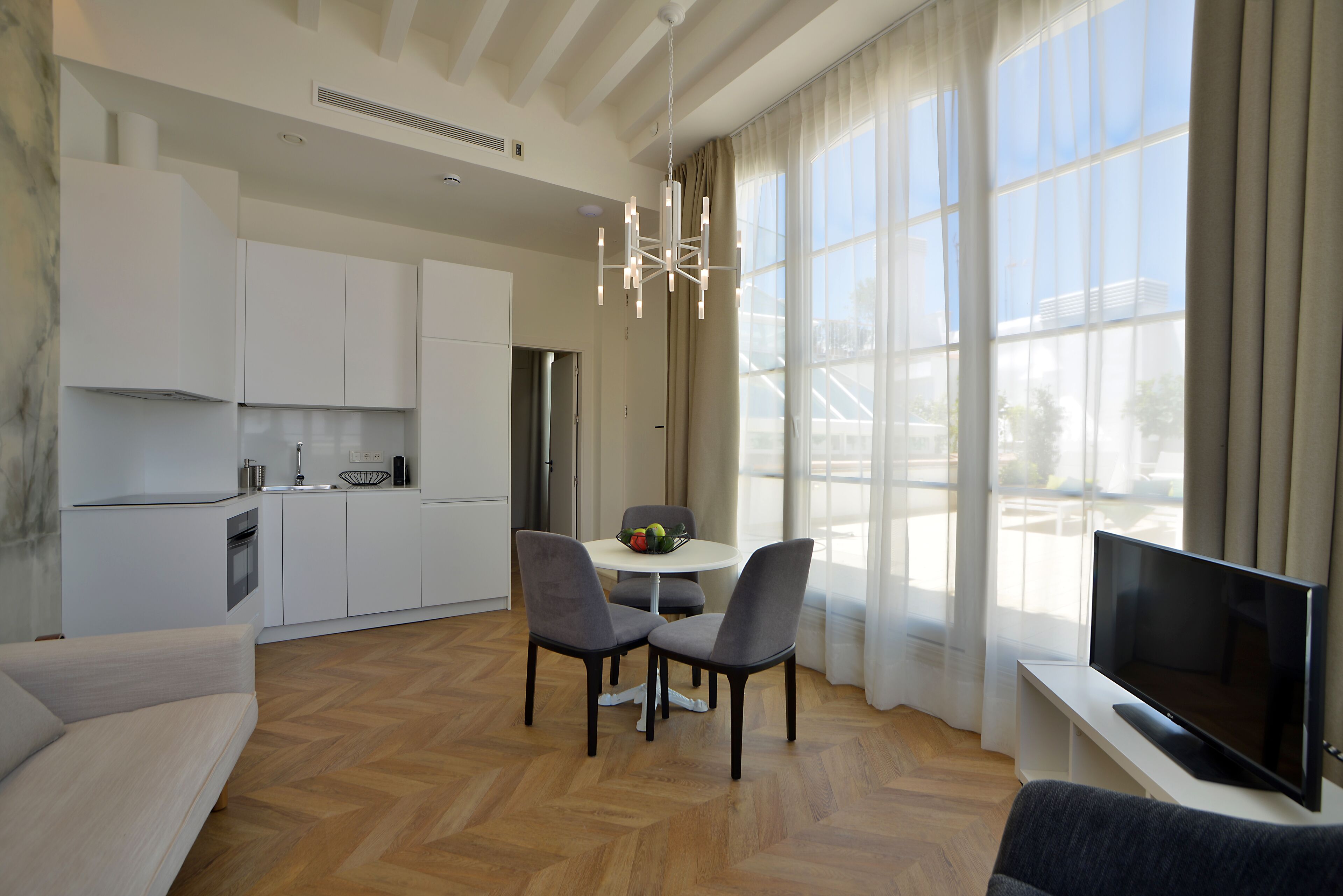 Uma sala de estar moderna com sofá, mesa de jantar com cadeiras, televisão de tela plana e área de cozinha com armários e janela.