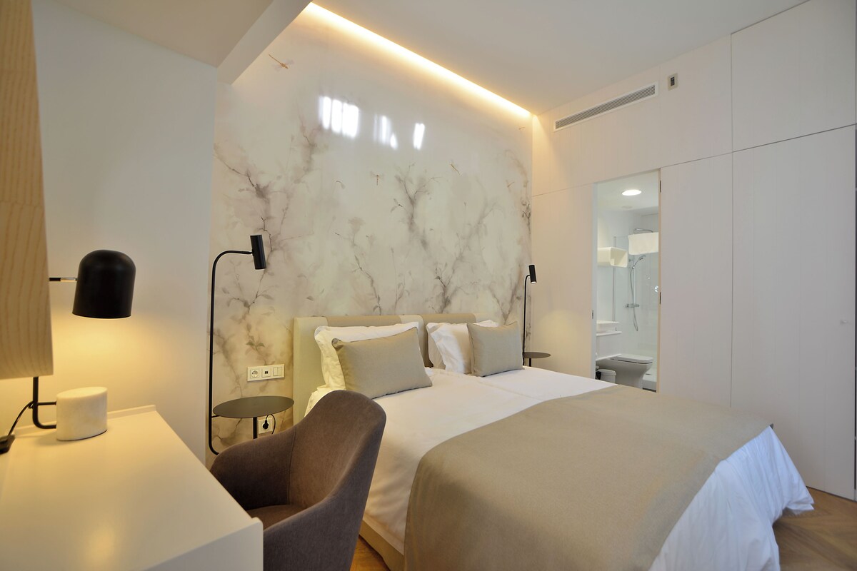 Quarto de hotel moderno com uma cama grande, uma cadeira, uma escrivaninha e um espelho.