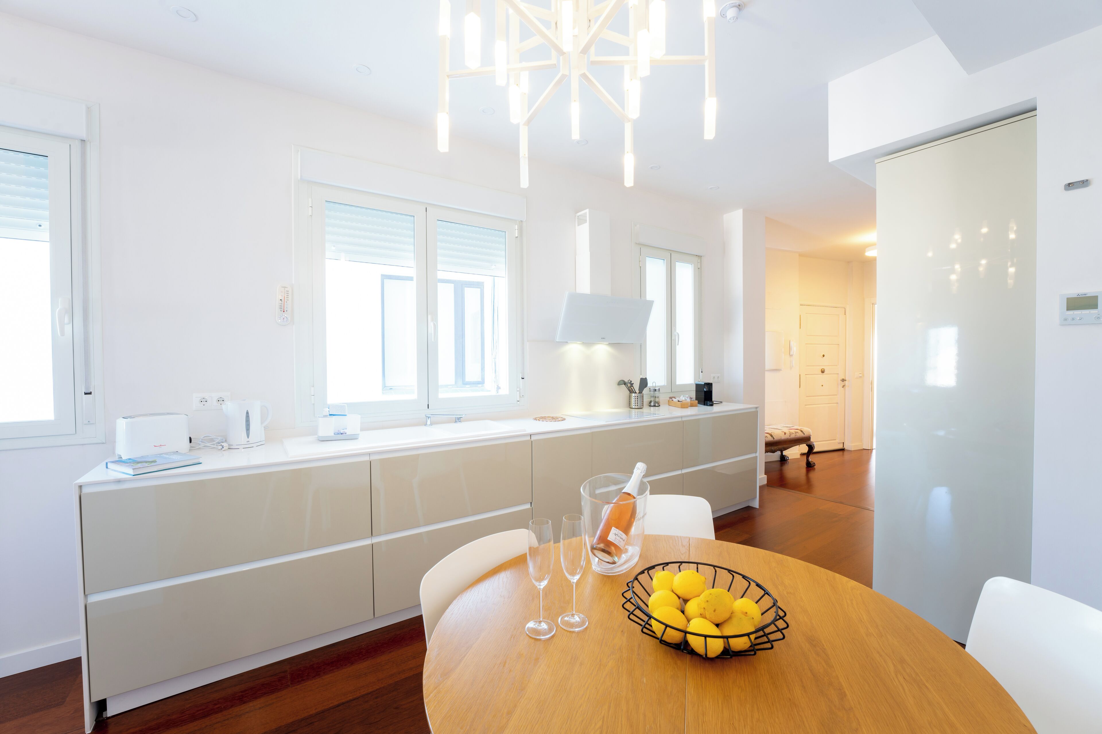 A modern kitchen with white cabinetry, a dining table with a fruit bowl, and a large window providing natural light.