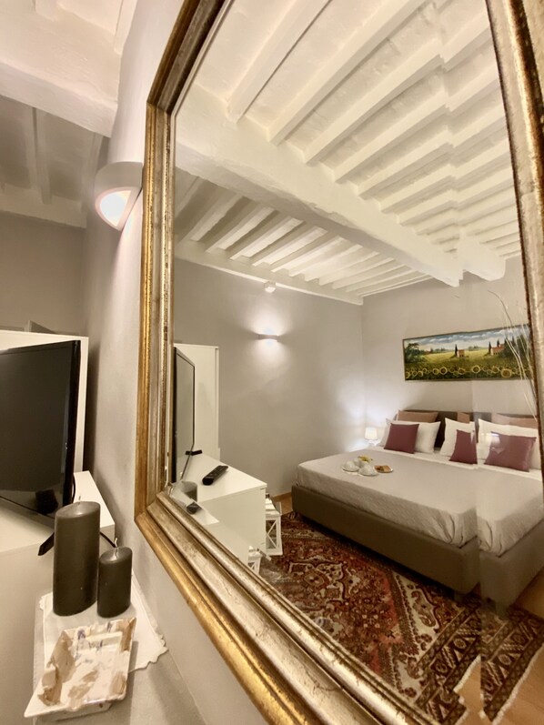 A hotel room with a large mirror reflecting the ceiling, a bed with pillows, a bedside table with a lamp, and a TV on a stand.