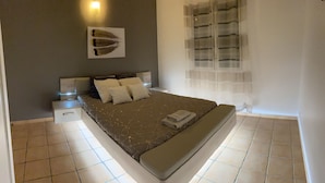 A bedroom with a bed, a nightstand, a wall-mounted artwork, a window with sheer curtains, and a tiled floor. - Villa Samathan 3 bedrooms private pool 5mn from the beach (Saint-François)
