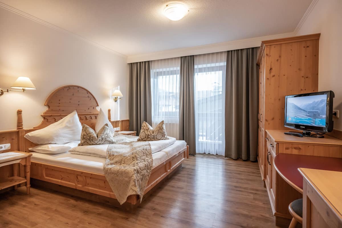 Una habitación de hotel con una cama de madera, un escritorio con televisor y un ventanal con cortinas.