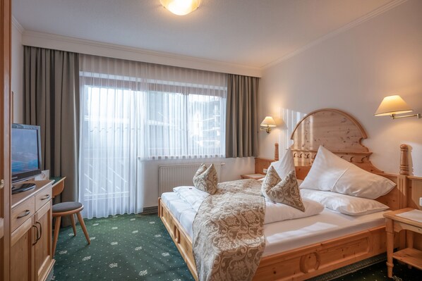 A hotel room with a wooden bed, a desk with a television, and a window with curtains. - Apparthotel Ederfeld (Mayrhofen)