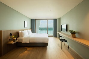 A modern hotel room with a large bed, a TV, a small table with a plant, and a view of the sea through the window.