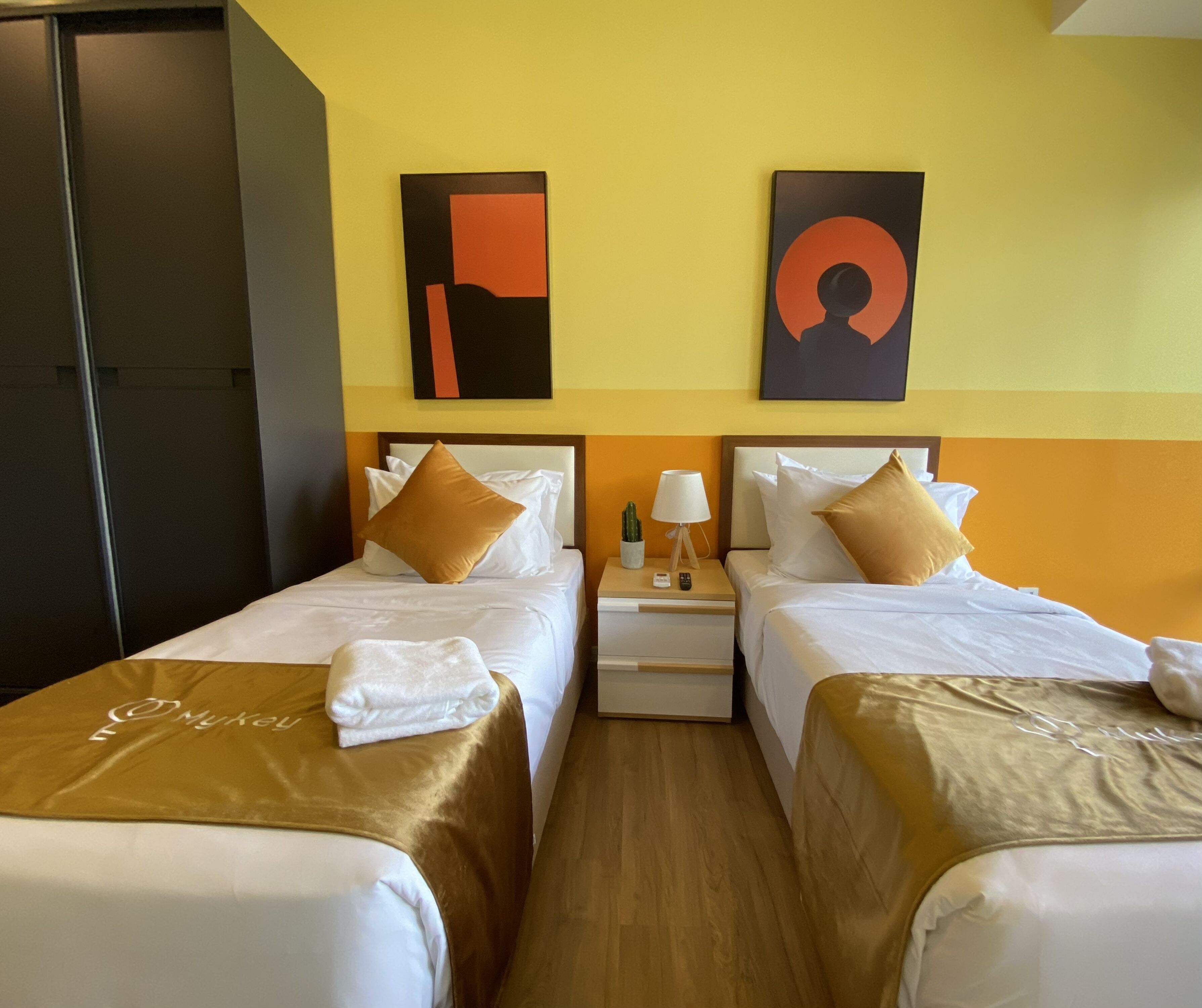 A hotel room with two beds, each with a brown headboard, white bedding, and a towel on the bed. There is a nightstand with a lamp and a cactus between the beds. The room has a yellow and orange wall color scheme.