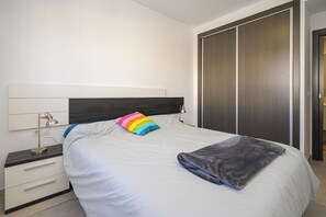 Een slaapkamer met een bed, een nachtkastje, een lamp, een kledingkast en een regenboogkleurig kussen.