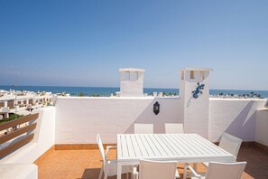 Een terras met een witte tafel en stoelen, uitzicht op zee, en een gebouw met een muurschildering.