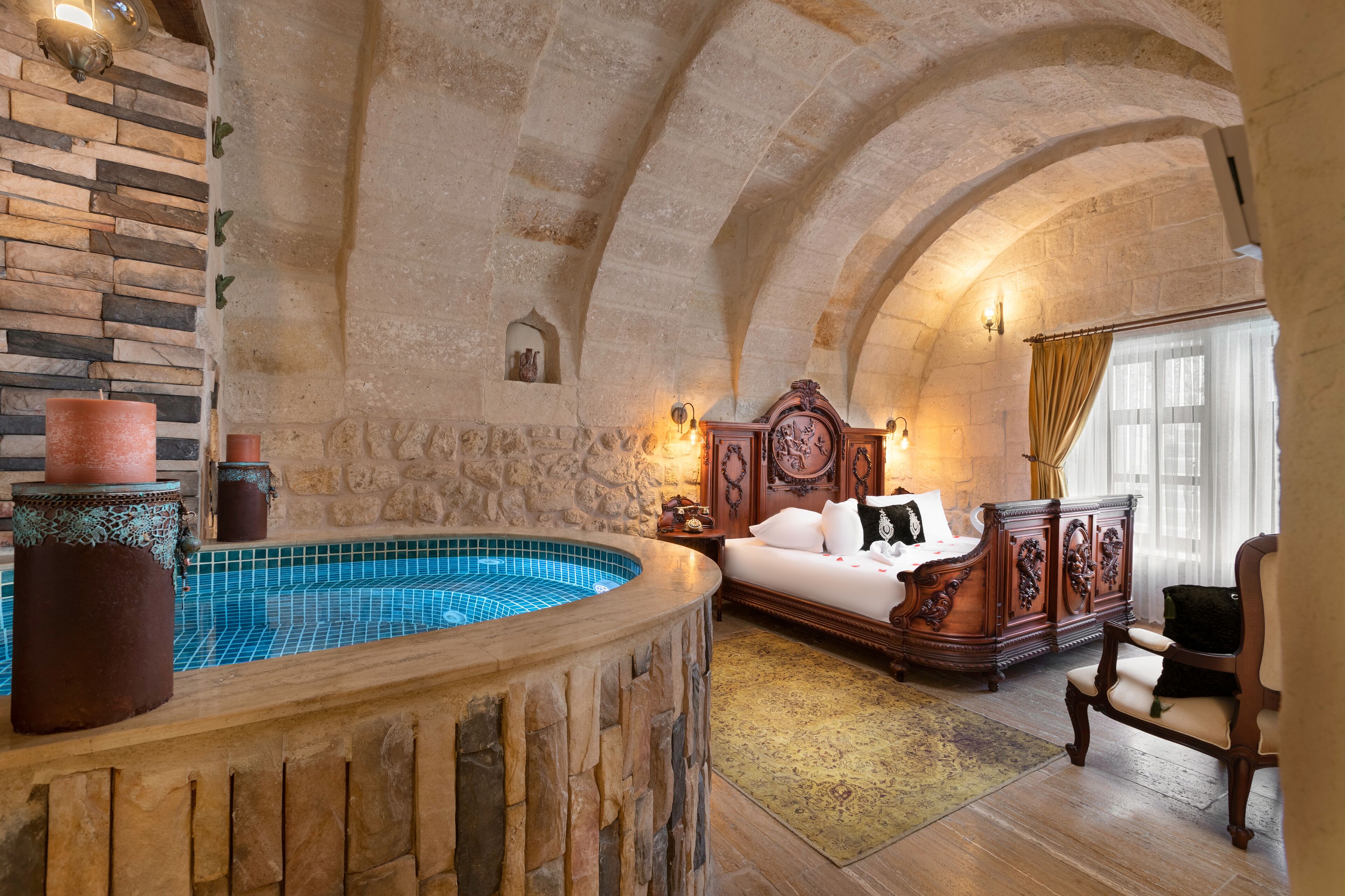 Una habitación con pared de piedra, una bañera de madera, una cama con cabecera, un aparador de madera y una silla.