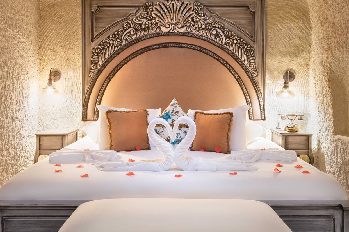 A luxurious hotel room with an ornate headboard, a neatly made bed with towels and rose petals, and two wall sconces.