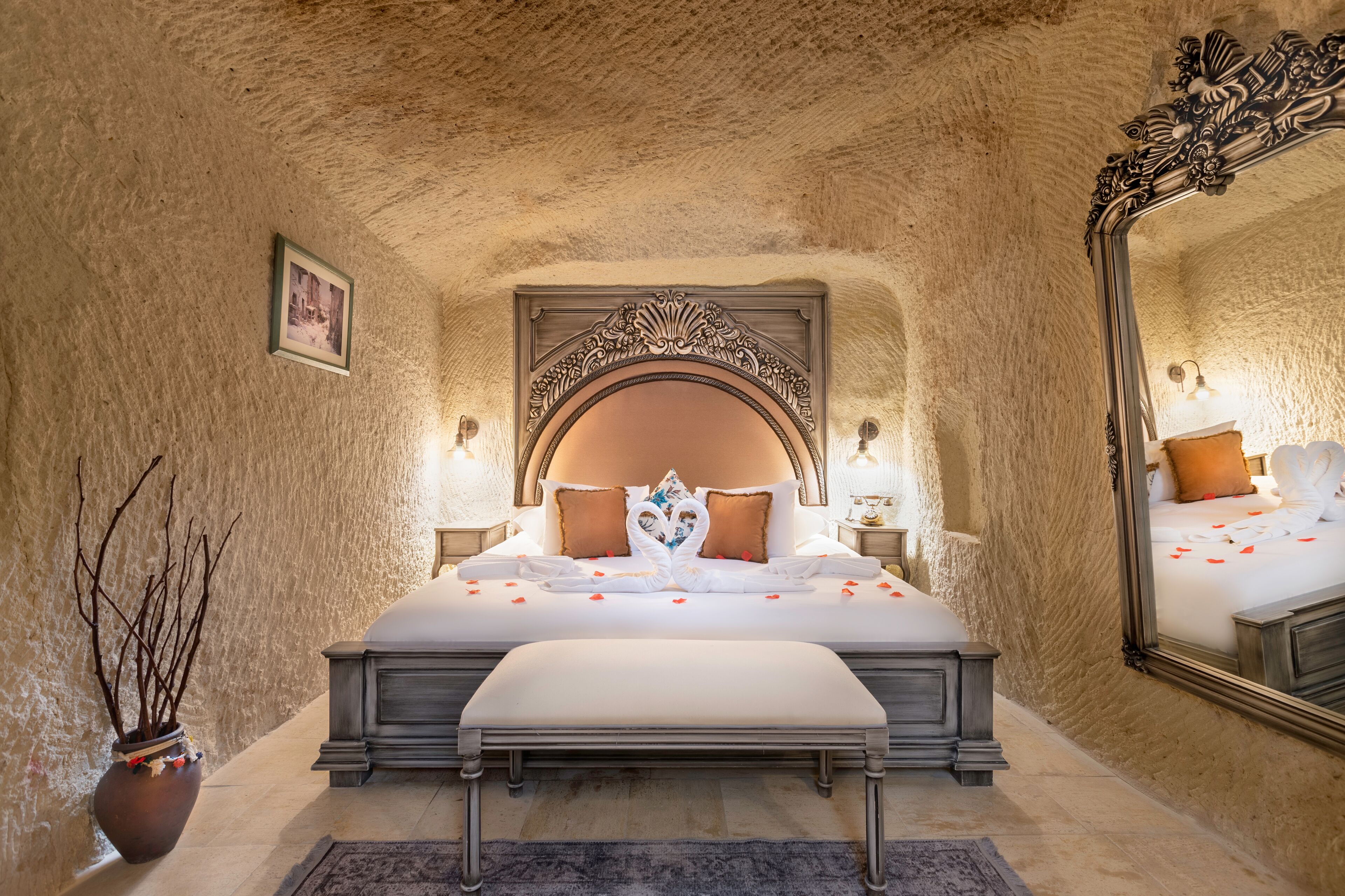 A bedroom with a large bed, ornate headboard, bedside lamps, a mirror, and a vase with branches.