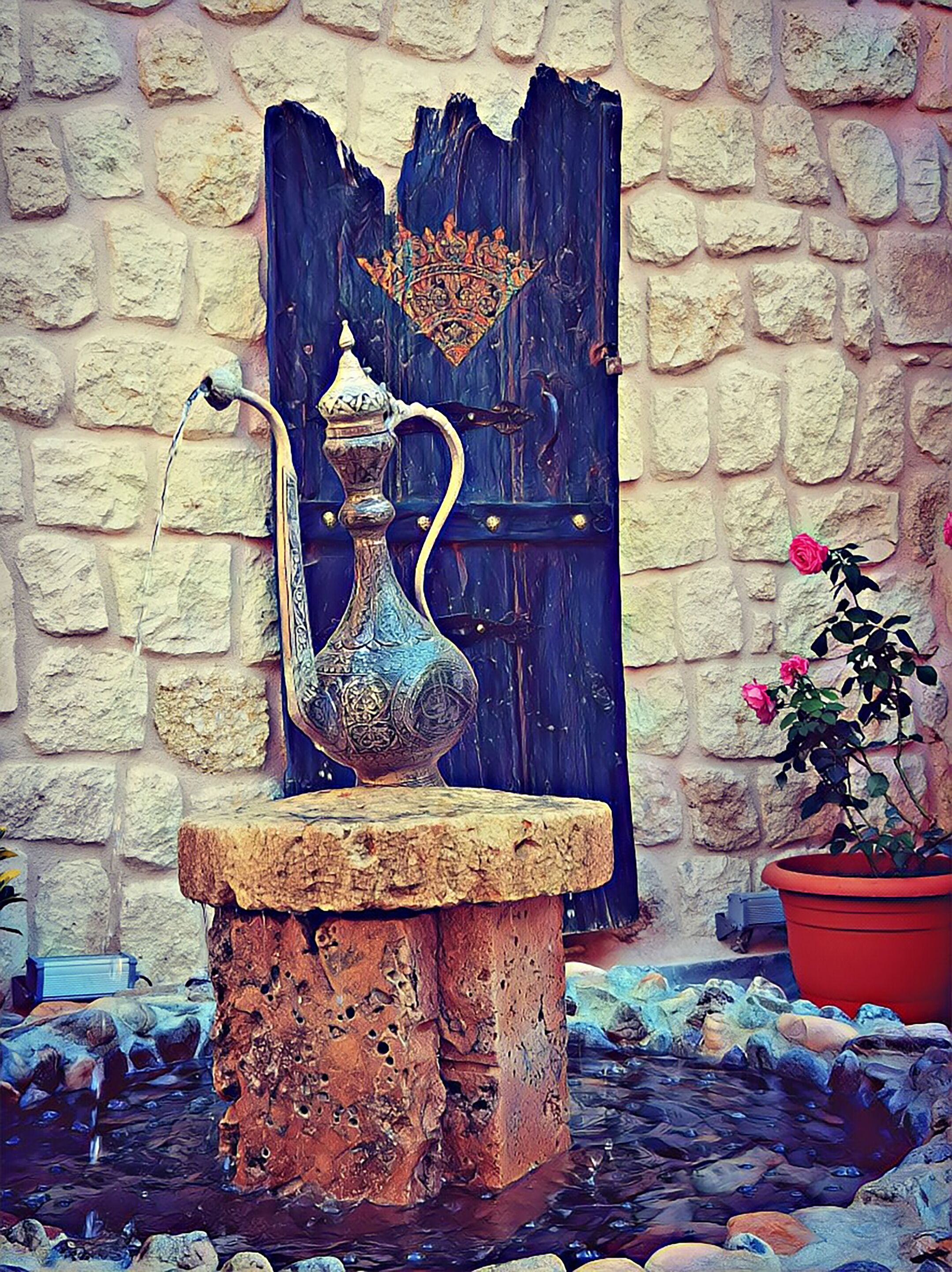 A stone fountain with a metal spout and a decorative jug, placed on a stone pedestal in front of a rustic stone wall and a wooden door.