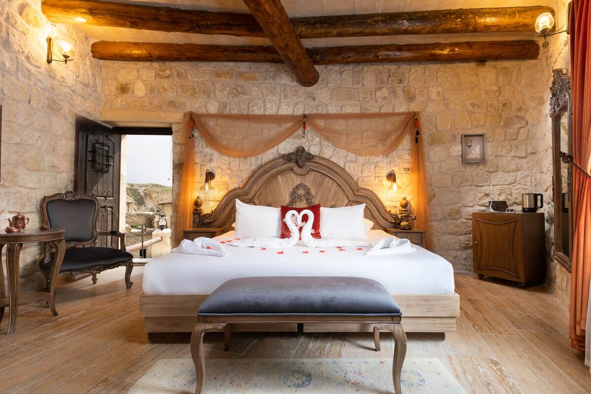 A bedroom with a stone wall, wooden beams, a bed with white linens, a bench, a chair, a small table, and a nightstand with a lamp.