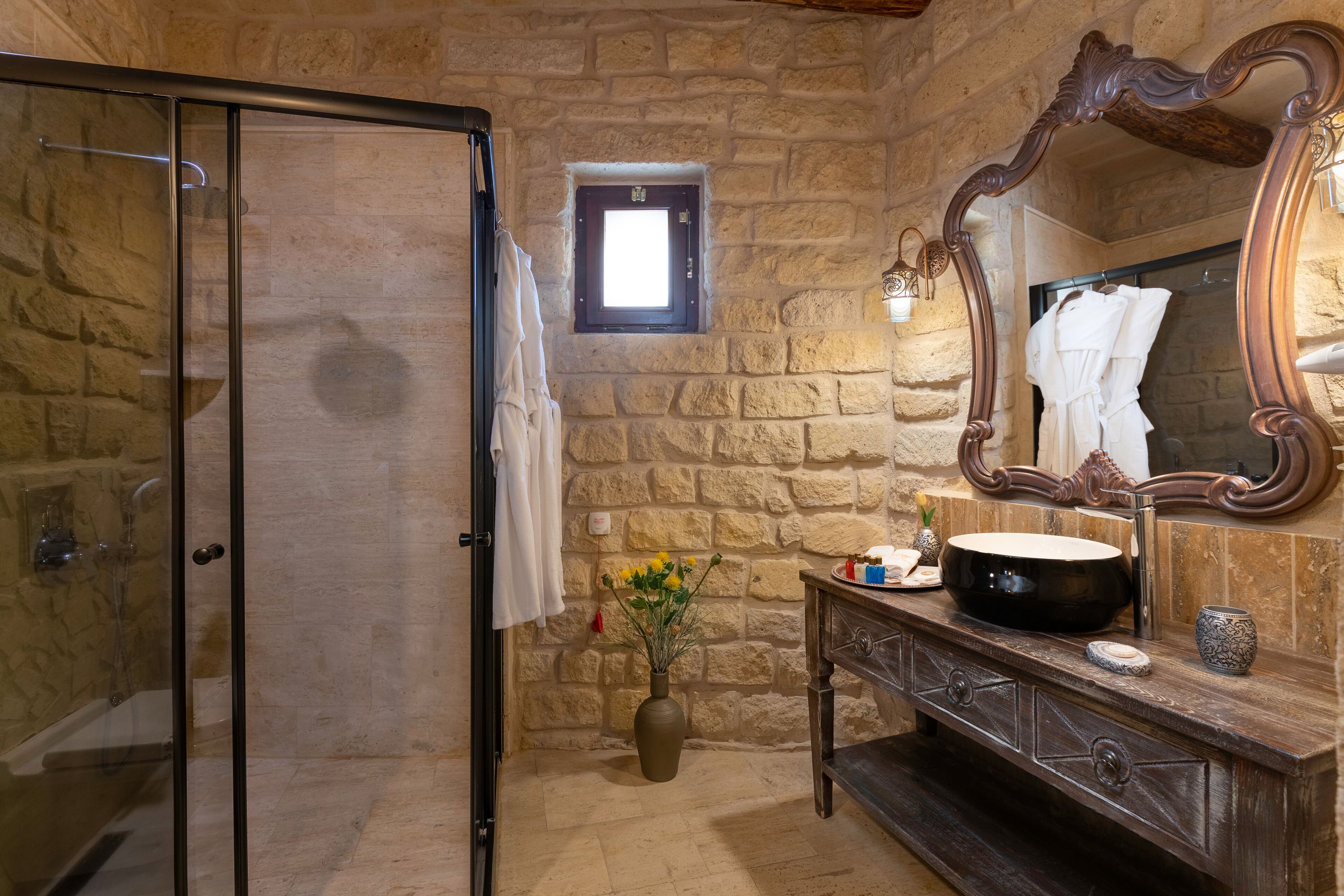 A bathroom with a stone wall, a glass shower enclosure, a wooden vanity with a large mirror, and a vase with flowers.