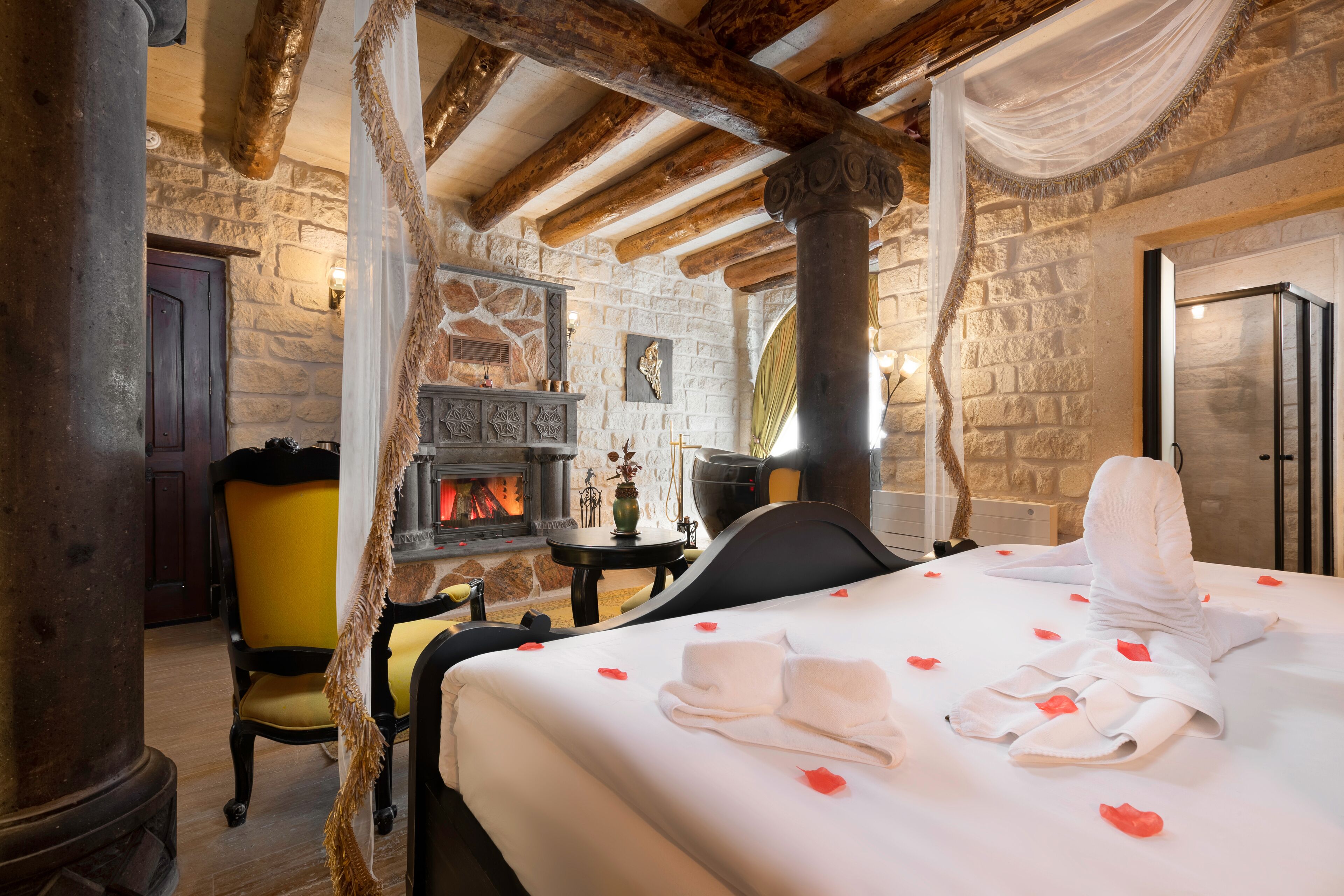 Habitación de hotel con pared de piedra, chimenea, cama con pétalos de rosa y una silla amarilla.