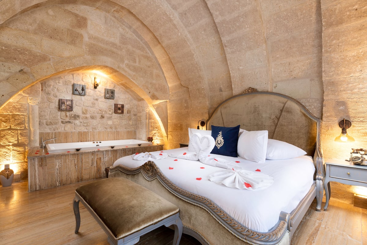 A bedroom with a stone arch, a bed with white linens, a bench, a bedside table, and a small sink.