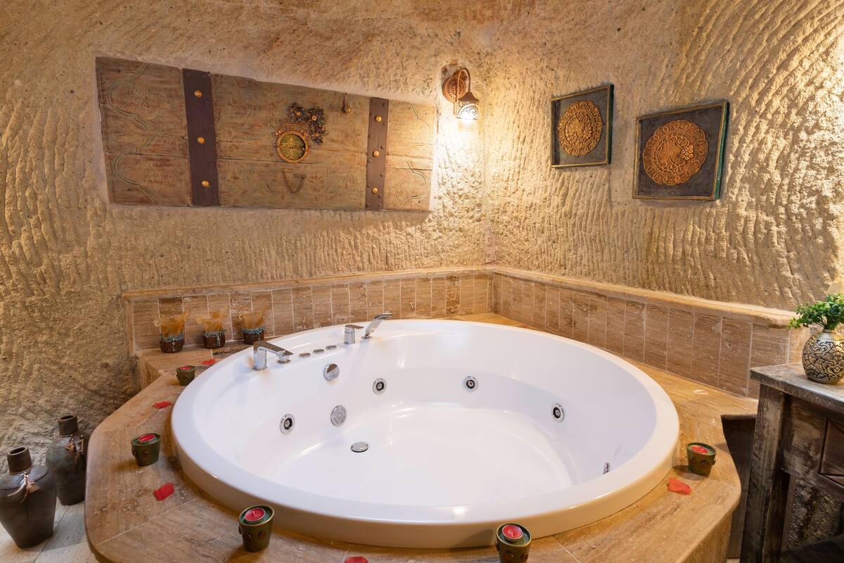 A large oval bathtub with multiple faucets, surrounded by a stone wall with decorative elements and a small plant on the right.