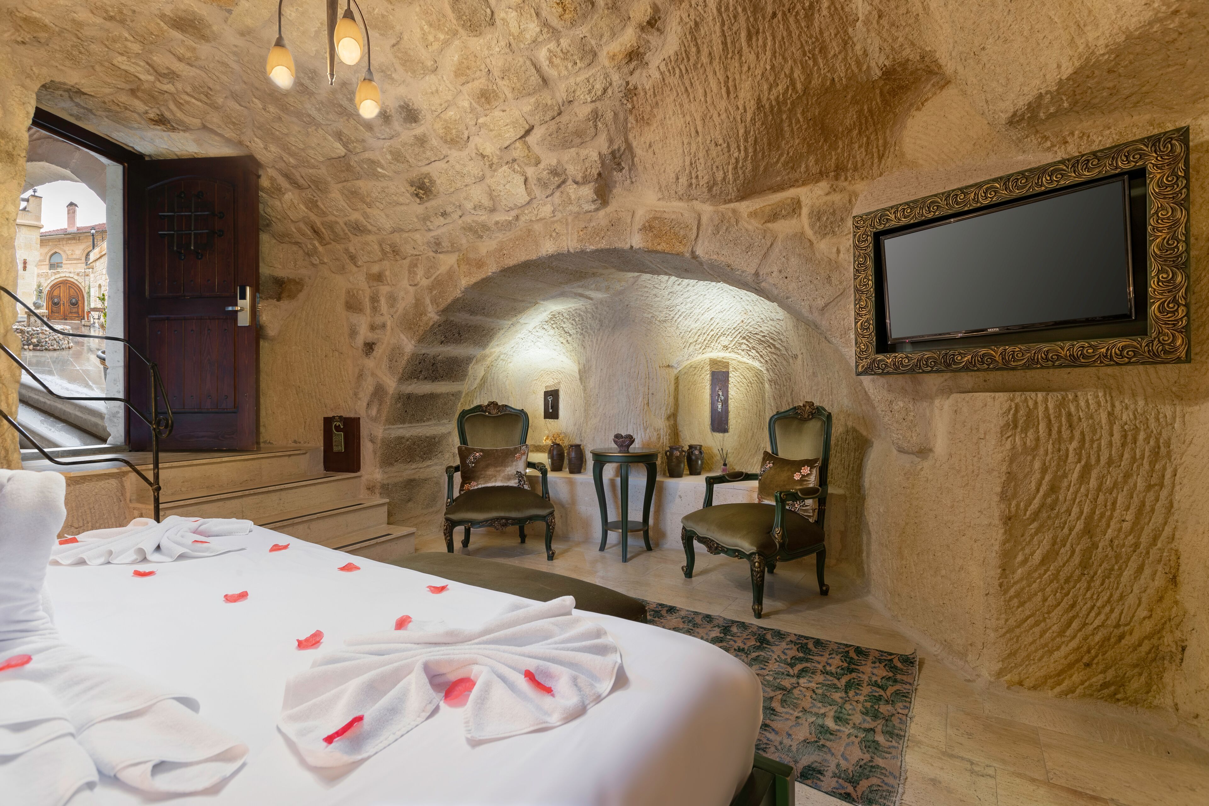 A hotel room with a stone archway, a bed with rose petals, two chairs, a small table, and a TV on the wall.