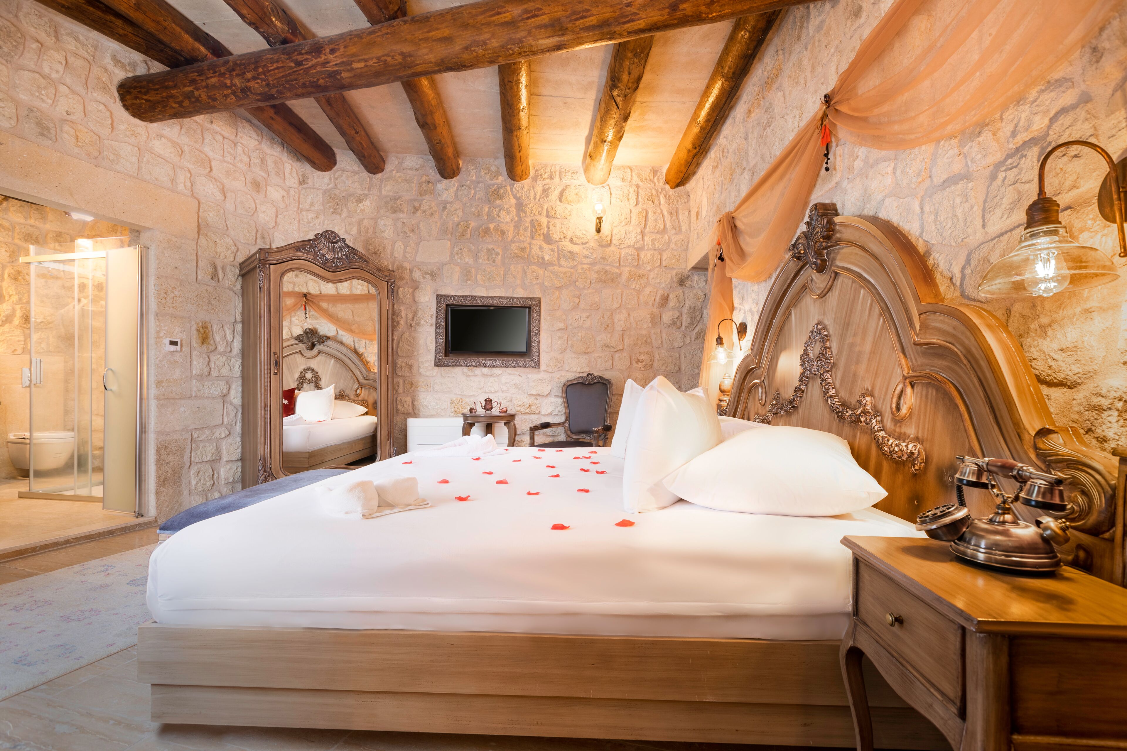 A bedroom with a stone wall, wooden ceiling beams, a large bed with white linens, a wooden nightstand, a mirror, and a flat-screen TV.