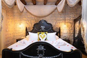 Una cama con cabecera oscura, ropa de cama blanca y almohadas con forma de corazón. Hay dos lámparas de noche y un espejo en la pared.