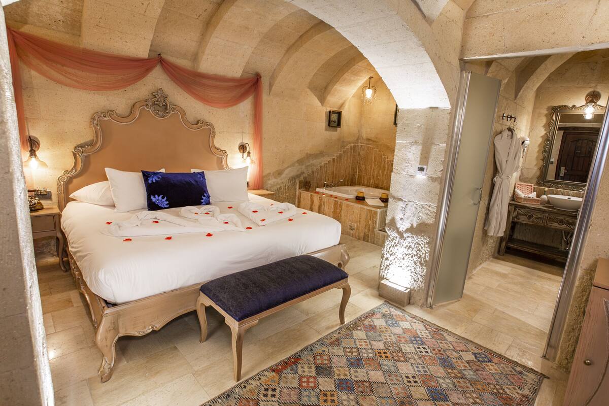 A bedroom with a large bed, a bench, a bathroom with a bathtub, and a shower area.