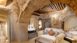 Un interior rústico con techo de piedra, un sofá blanco con cojines florales, una mesa de centro de madera y una cama con cabecera de madera.