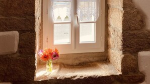 Una pequeña ventana con cortinas de encaje, una viga de madera encima y un florero con flores en el alféizar.