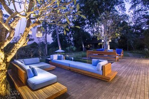An outdoor deck with wooden furniture and a glass-top table, surrounded by trees and a small water feature. - Villa Alam Taru (Cemagi)