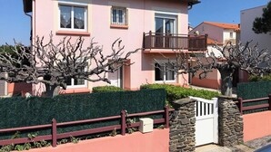 A pink two-story house with a balcony, surrounded by hedges and a stone wall. - Spacious Villa for 10, 100m from Beach, WiFi, Garden, A/C, Near Shops, Parking Available (Le Barcarès)