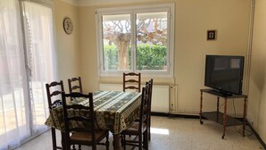A dining area with a table set for four, a television on a stand, and a window with a view of greenery. - Spacious Villa for 10, 100m from Beach, WiFi, Garden, A/C, Near Shops, Parking Available (Le Barcarès)