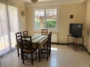 A dining area with a table set for four, a television on a stand, and a window with a view of greenery.