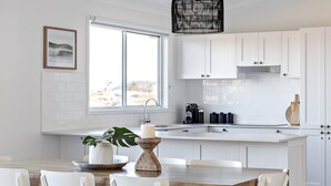 A modern kitchen with white cabinetry, a large window, and a dining area with a wooden table and chairs. - Ohana - Amazing Views (Gerroa)