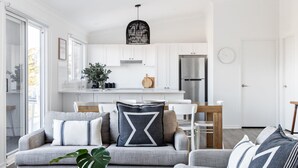 A modern kitchen with white cabinets, a stainless steel refrigerator, and a dining area with a wooden table and white chairs. - Ohana - Amazing Views (Gerroa)