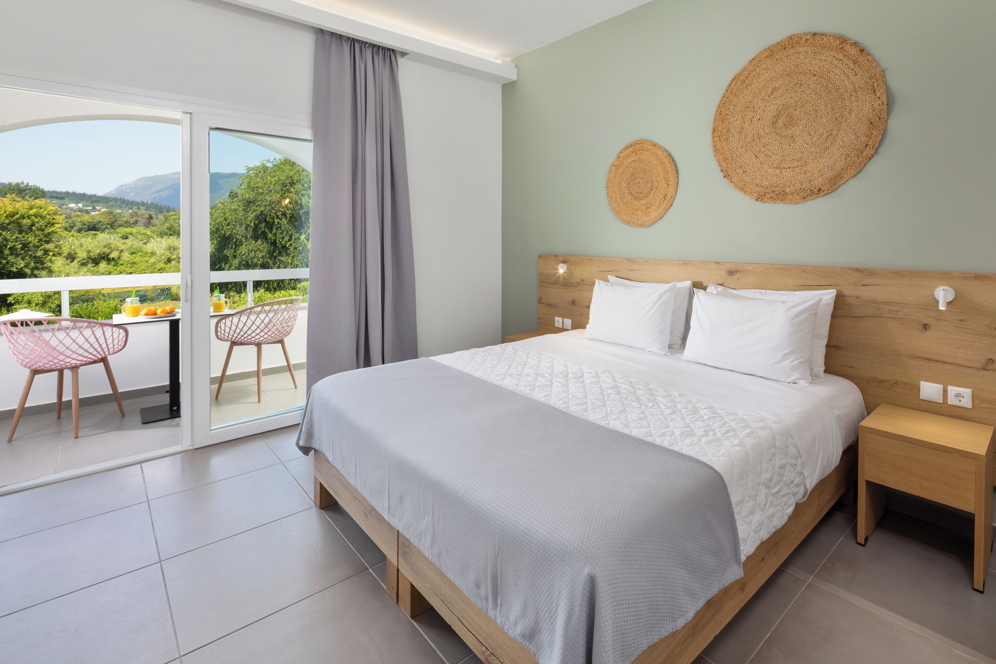 A bedroom with a large bed, a nightstand, a wooden headboard, two woven wall decorations, and a balcony with chairs and a table.