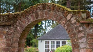 An arched brick gateway leading to a white house with a dark roof, surrounded by lush greenery and flowering plants. - Broad House  - sleeps 8 guests  in 4 bedrooms (Dilham)