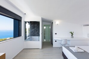 Enjoy a serene sea view from the large window, may relax in the separate shower area, and rest on the neatly made bed with a patterned bedspread.