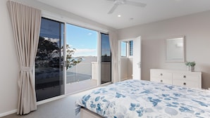 A bedroom with a large sliding glass door leading to a balcony, a bed with a floral bedspread, a white dresser, and a ceiling fan. - Iluka, 46 Soldiers Point Rd - Stunning Air Conditioned house with WIFI & Water Views (Soldiers Point)