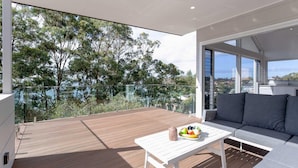 A balcony with a wooden deck, a white table with a fruit bowl, and a grey sofa. - Iluka, 46 Soldiers Point Rd - Stunning Air Conditioned house with WIFI & Water Views (Soldiers Point)