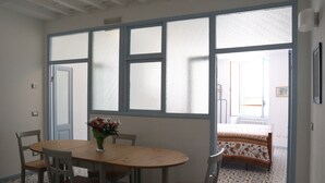 A dining area with a wooden table and chairs, a vase with flowers, and a view through frosted windows to a bedroom with a patterned bedspread.