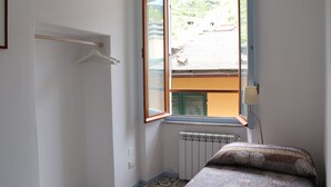 A small bedroom with a single bed, a radiator, a window with a view of a building, and a wall-mounted light fixture.