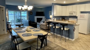 Una cocina moderna con armarios blancos, una isla central con taburetes de bar, un comedor con mesa y sillas, y una sala de estar con sofá y chimenea.