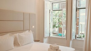 A hotel room with a bed, pillows, a nightstand, and a window with a view of the outside. - Le Premier Lisbon Suites (Lisbon)