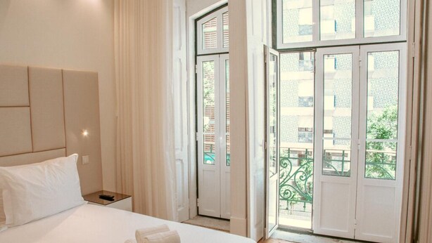 A hotel room with a bed, a nightstand, and a view of a balcony with a railing. - Le Premier Lisbon Suites (Lisbon)