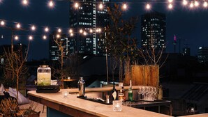 Un bar situé sur un toit, avec une vue imprenable sur les toits de la ville, des guirlandes lumineuses et divers équipements typiques d’un bar.