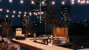 A rooftop bar with a city skyline in the background, string lights, and various bar amenities. - Ruby Louise Hotel Frankfurt by IHG (Frankfurt)